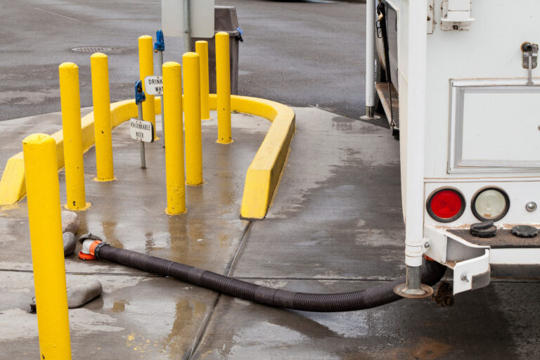 RV at a dump station connected to a sewer hose, with non-potable and potable water spigots visible and surrounded by yellow safety posts on a wet concrete pad.